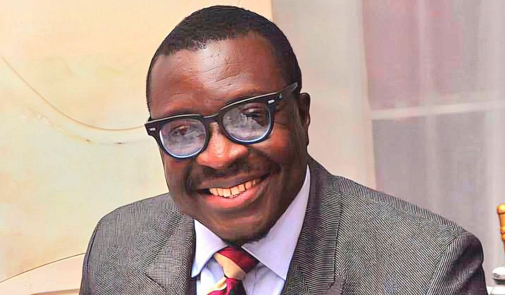 Comedian Ali Baba seated, wearing a gray suit with a patterned design, a white shirt, and a red tie with diagonal stripes.