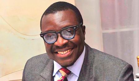 Comedian Ali Baba seated, wearing a gray suit with a patterned design, a white shirt, and a red tie with diagonal stripes.