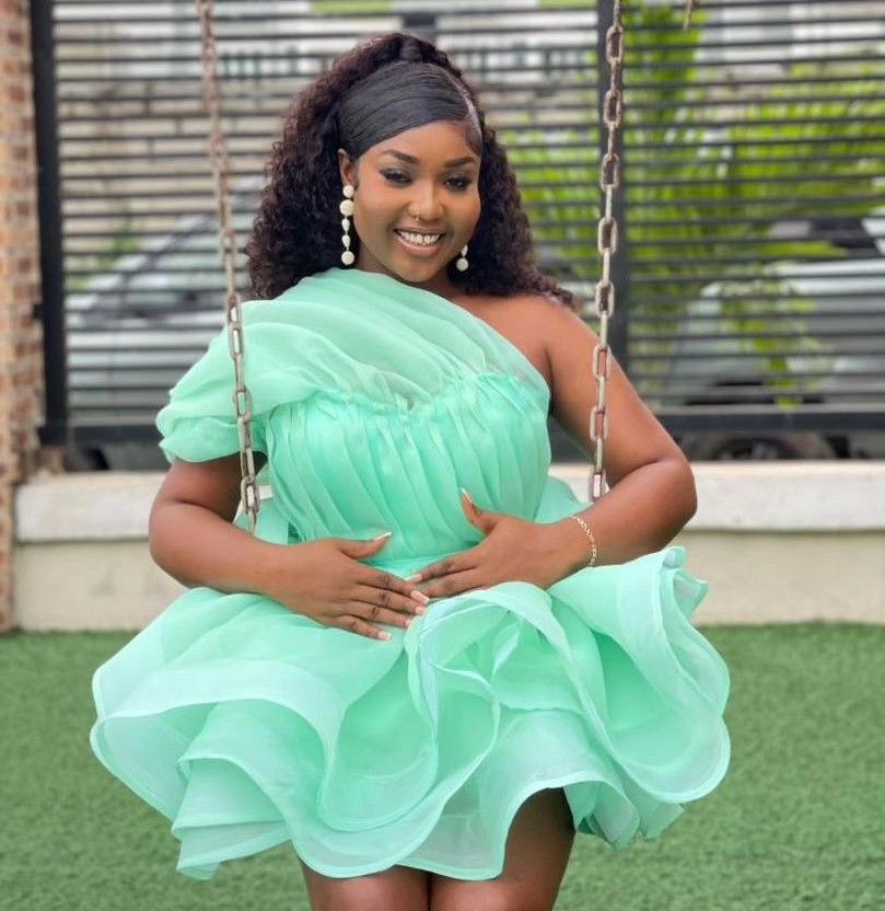 BBNaija star Rhuthee seated on a swing, wearing a mint green, off-the-shoulder ruffle dress. Her long, dark curly hair cascades down her back as she sits in a relaxed posture with her hands gently placed on her lap. The background features a blurred view of an outdoor garden setting, adding to the serene and stylish ambiance.
