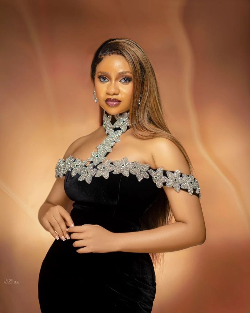 Fashion portrait of BBNaija star Victoria in an elegant black dress with silver floral accents, set against a warm-toned backdrop.