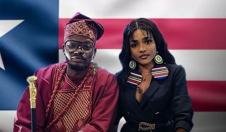 BBNaija couple Kellyrae and Kassia seated together. Kellyrae on the left is wearing a traditional red and gold patterned outfit with a matching hat and holding a cane or staff. Kassia on the right is dressed in a black outfit with colorful stripes at the collar and large circular earrings. In the background, there is a flag resembling the Liberian flag.