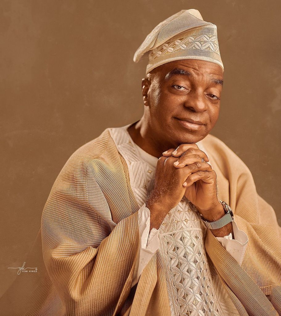 Bishop David Oyedepo, dressed in a cream-coloured agbada with intricate embroidery and a matching cap, seated with hands clasped together in a pose of reflection.