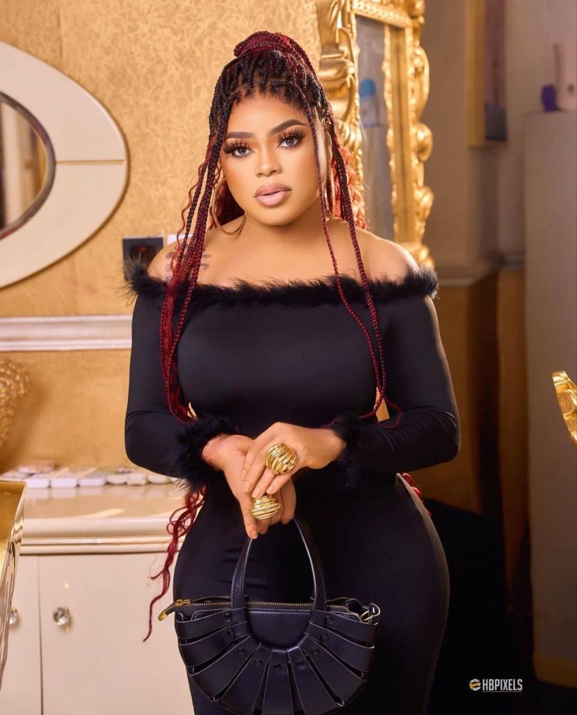 Bobrisky, dressed in an elegant black outfit, posing indoors with braided red and black hair and holding a black handbag.