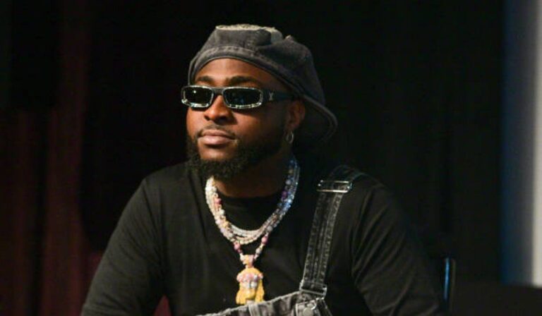 Afropop singer Davido with an obscured face wearing a black shirt, multiple necklaces, and a hat, sitting in front of a blurred background.