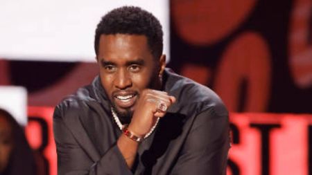 Diddy seated at an event, wearing a black outfit with a red bracelet, making a gesture with his hand near his chin. The background features red tones and lighting