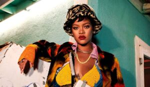 Rihanna wears a stunning, floor-length red gown with intricate detailing. Her hair is styled in loose waves, and she accessorizes with elegant jewelry, including statement earrings and a matching bracelet. She exudes confidence and glamour, making a striking appearance at the event.