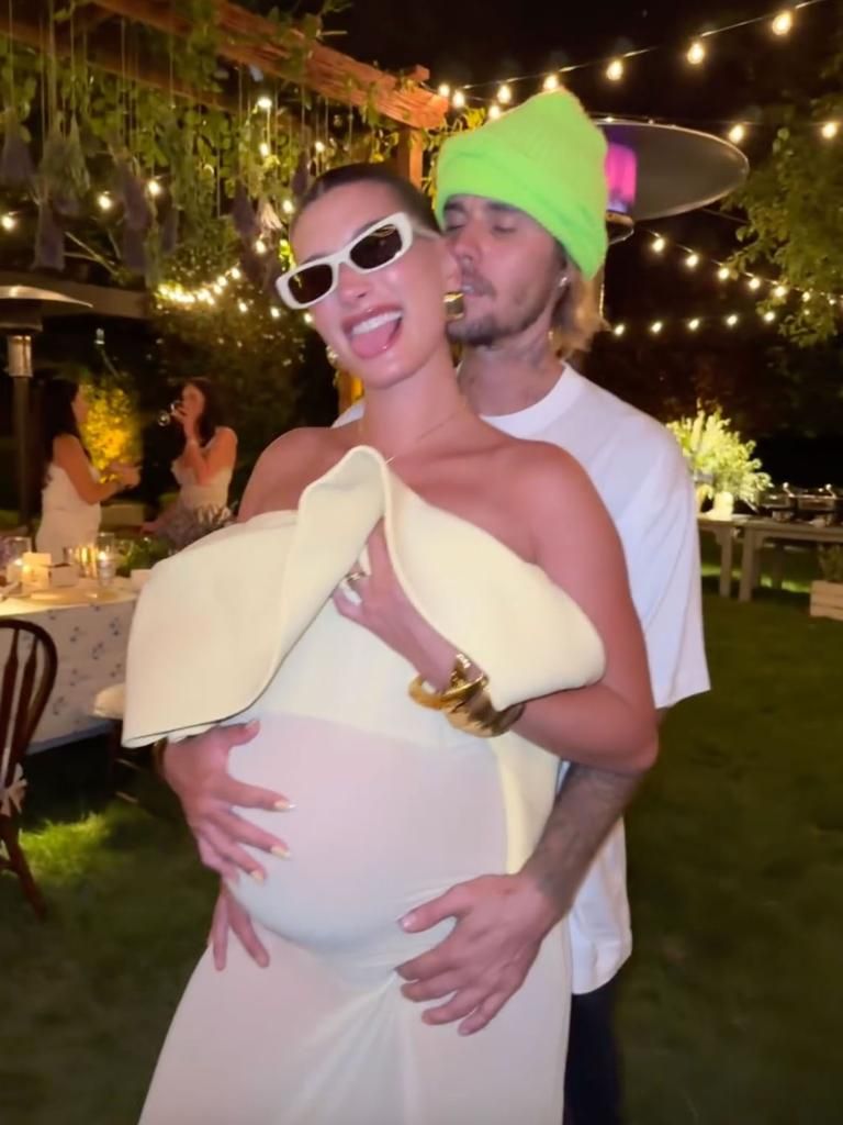 Justin Bieber and his wife Hailey. Justin has on a green cap and white sleeveless top, and Hailey is wearing an off-shoulder white dress or top.