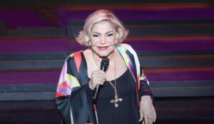 Legendary Greek singer Marinella performing on stage, holding a microphone and wearing a black top with colorful patterns.