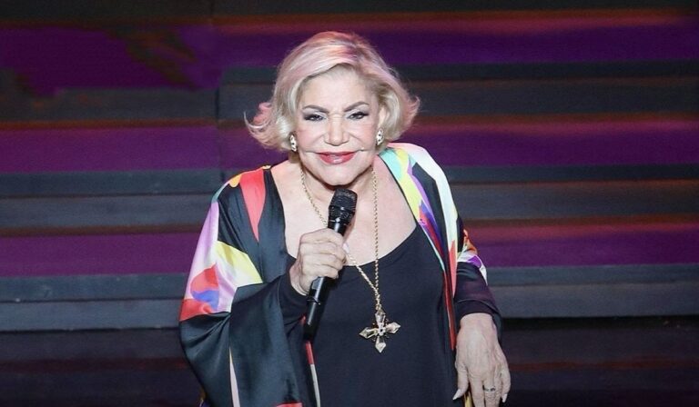 Legendary Greek singer Marinella performing on stage, holding a microphone and wearing a black top with colorful patterns.