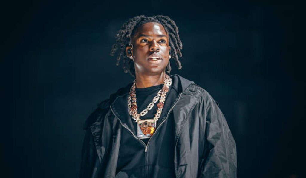 Rema standing on stage, from the chest up, wearing a black jacket and multiple chain necklaces with a pendant.