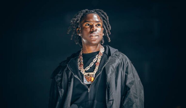 Rema standing on stage, from the chest up, wearing a black jacket and multiple chain necklaces with a pendant.