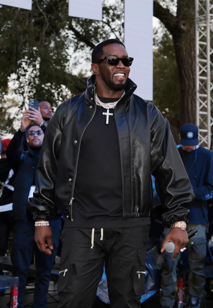 Sean “Diddy” Combs, dressed in a black jacket and pants with white drawstrings, wearing multiple pieces of jewelry including a cross pendant necklace, stands confidently with his hands partially in his pockets. The background features blurred figures, suggesting a crowd or gathering.