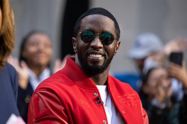Sean Diddy Combs wearing a red jacket with “Sean John” embroidered on it, sitting in front of blurred figures.