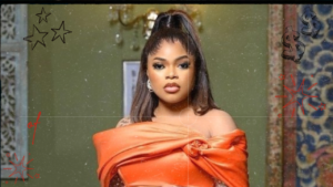Bobrisky draped in an elegant orange attire stands against a textured backdrop, exuding a sense of style and mystery.