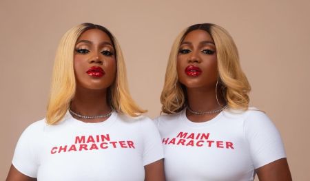 Twin sisters Wanni and Handi pose confidently, wearing matching white t-shirts that read "MAIN CHARACTER" in bold red letters. Their striking looks are complemented by glossy red lips and stylish blonde hair, set against a neutral background.