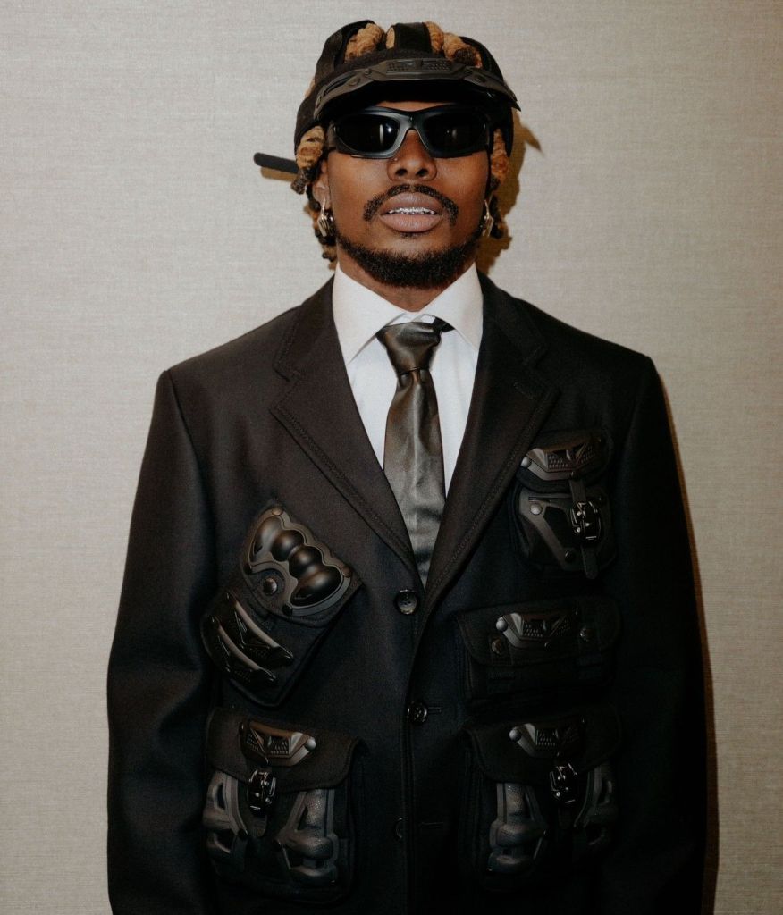 Asake poses confidently in a stylish outfit, featuring a black suit with multiple utility pockets and accessories, including sunglasses and a hat. The backdrop is simple and elegant, highlighting his bold fashion statement and unique flair.