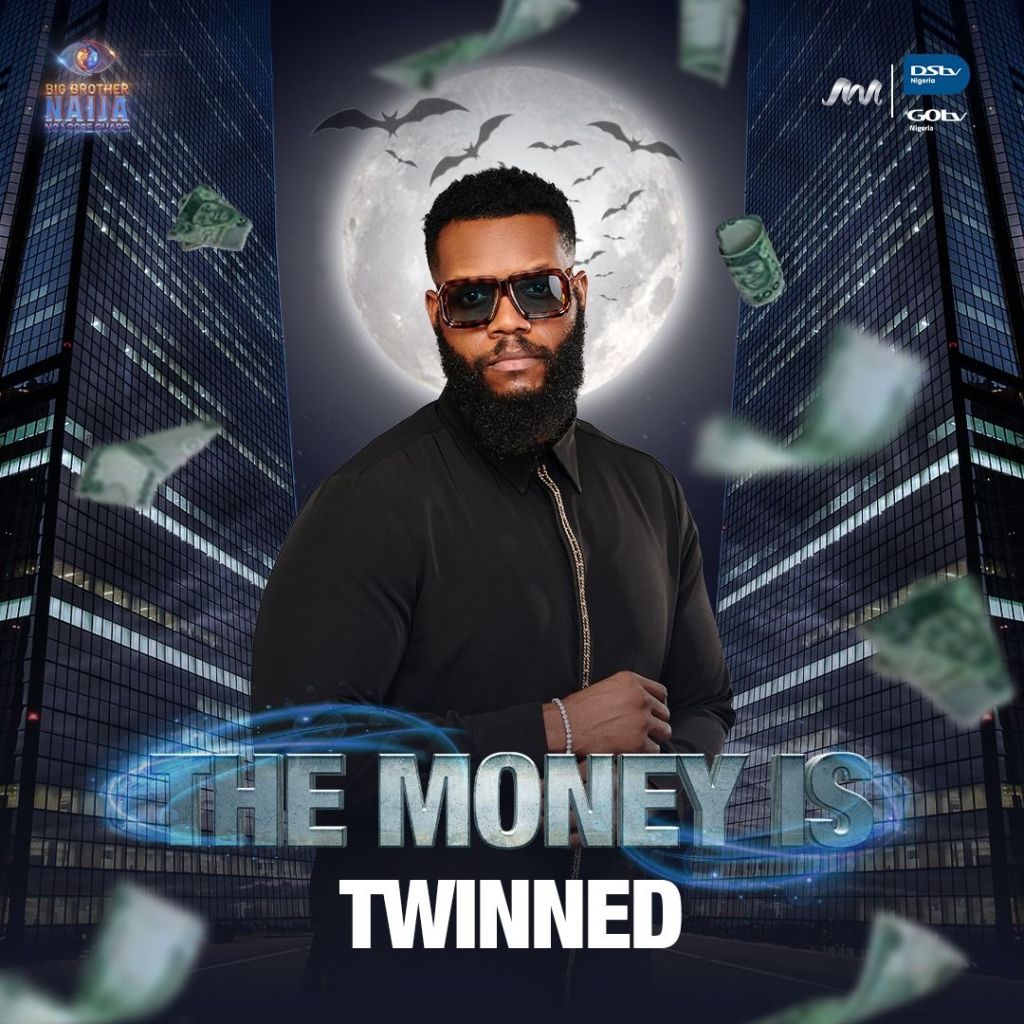 BBNaija star Ozee standing confidently in front of a dark cityscape, wearing sunglasses and a black shirt. A full moon and flying bats are visible in the background, along with scattered dollar bills and the text "THE MONEY IS TWINNED" prominently displayed.