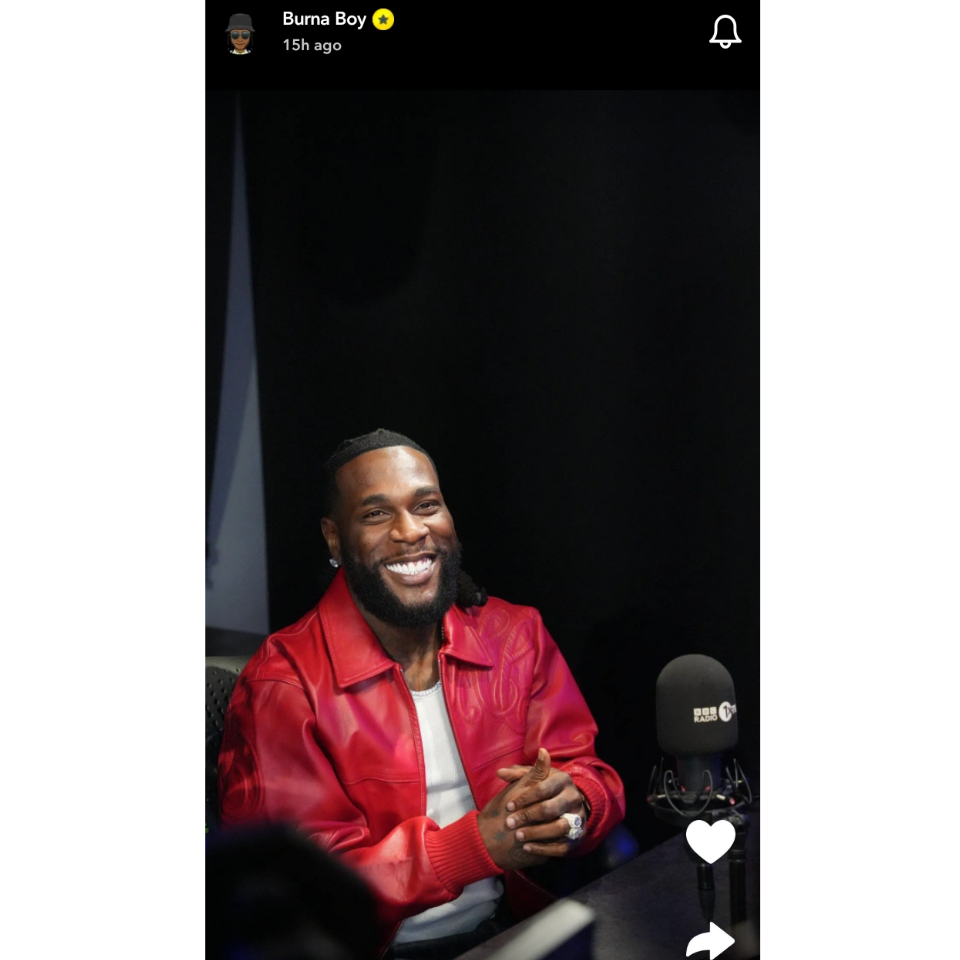 A smiling Burna Boy is seated in a recording studio, wearing a vibrant red jacket over a white shirt. The background is dark, highlighting his cheerful expression and stylish look, as he interacts with the microphone in front of him.