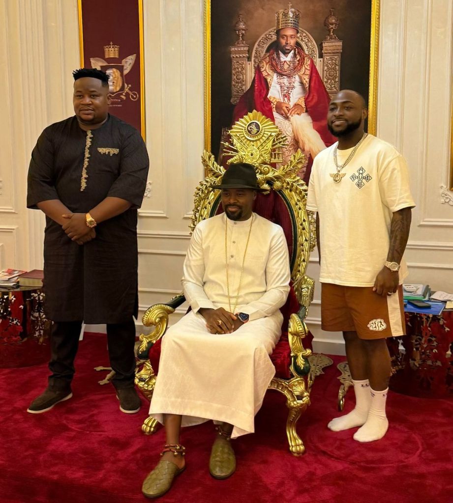 Cubana Chief Priest and singer Davido pose with His Royal Highness Olu of Warri, who is seated on an ornate throne. Chief Priest stands on the left in a black outfit, while Davido stands on the right in a casual white shirt and brown shorts, both smiling for the camera.