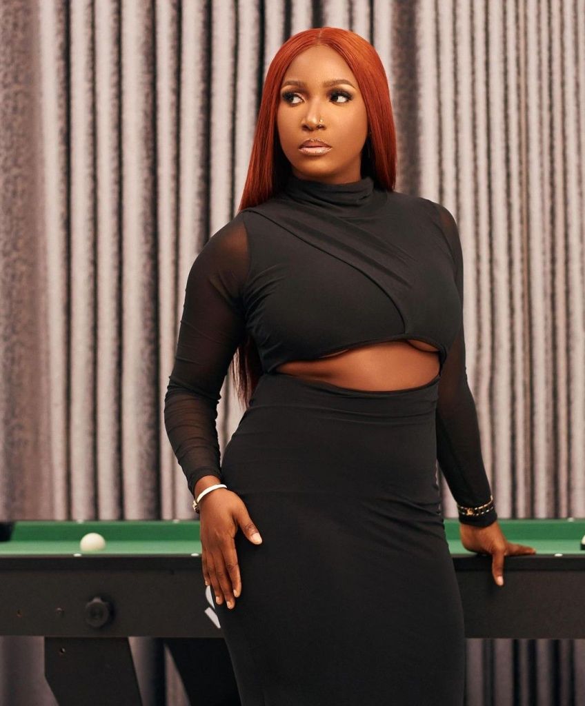 Damilola in a glamorous pose with sleek, straight red hair, showcasing her stylish look against a backdrop of textured curtains.