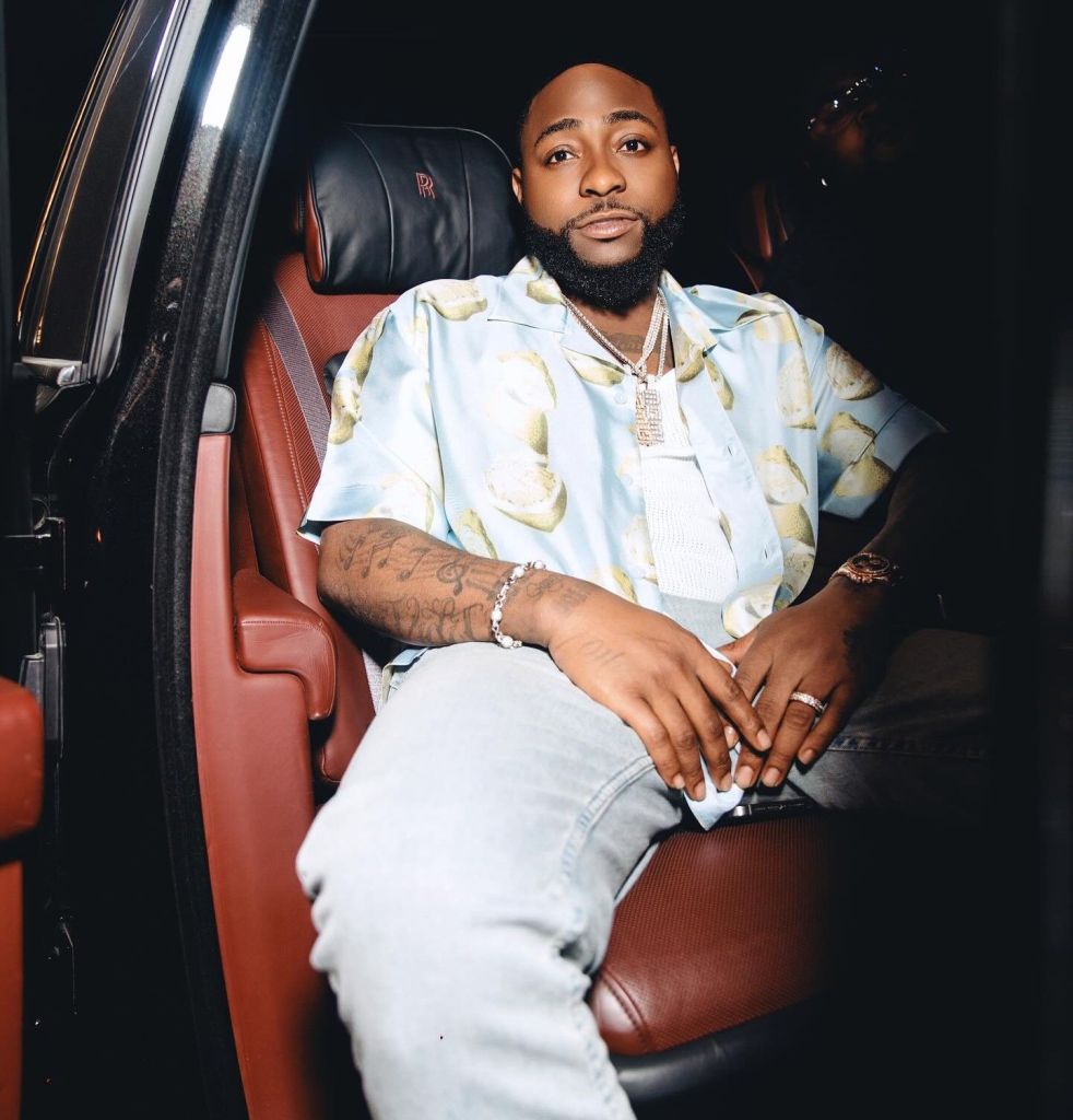 Davido in a colourful shirt with a necklace while sitting in a car.