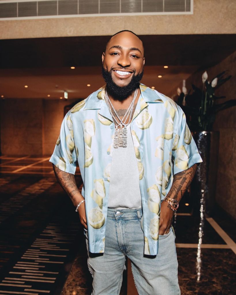 Davido smiles in a colourful patterned shirt, adorned with multiple chains and a warm demeanour.