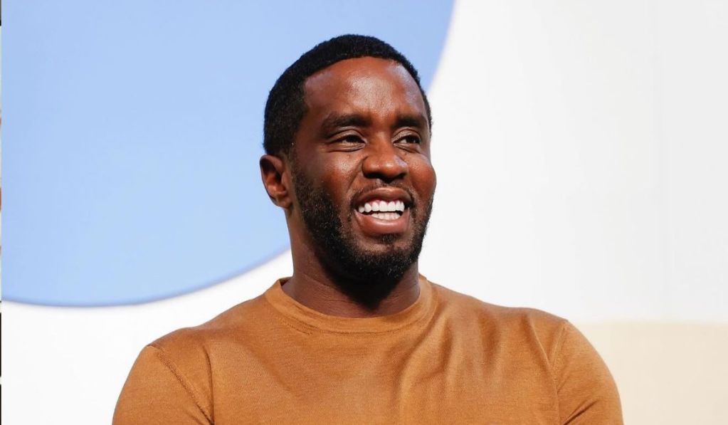 Diddy smiling and wearing a brown sweater is seated against a light backdrop, exuding a cheerful demeanor.