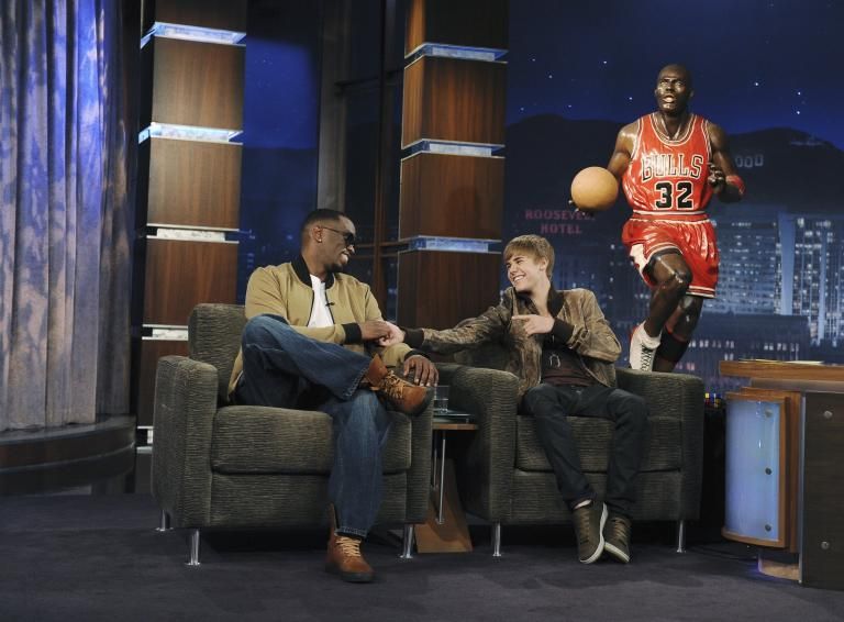 Justin Bieber and Sean "Diddy" Combs share a light-hearted moment during a talk show appearance, seated on a cozy set. Bieber is smiling and gesturing while Diddy laughs, and a cutout of Michael Jordan is displayed in the background.