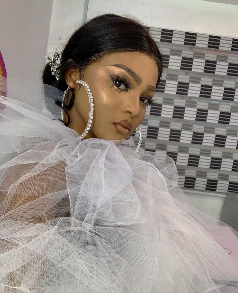 Kassia poses gracefully, draped in a voluminous white tulle outfit. She wears large, sparkling hoop earrings and showcases a radiant makeup look with bold brows and glossy lips. Her hair is styled elegantly, and she gazes confidently at the camera against a patterned backdrop.