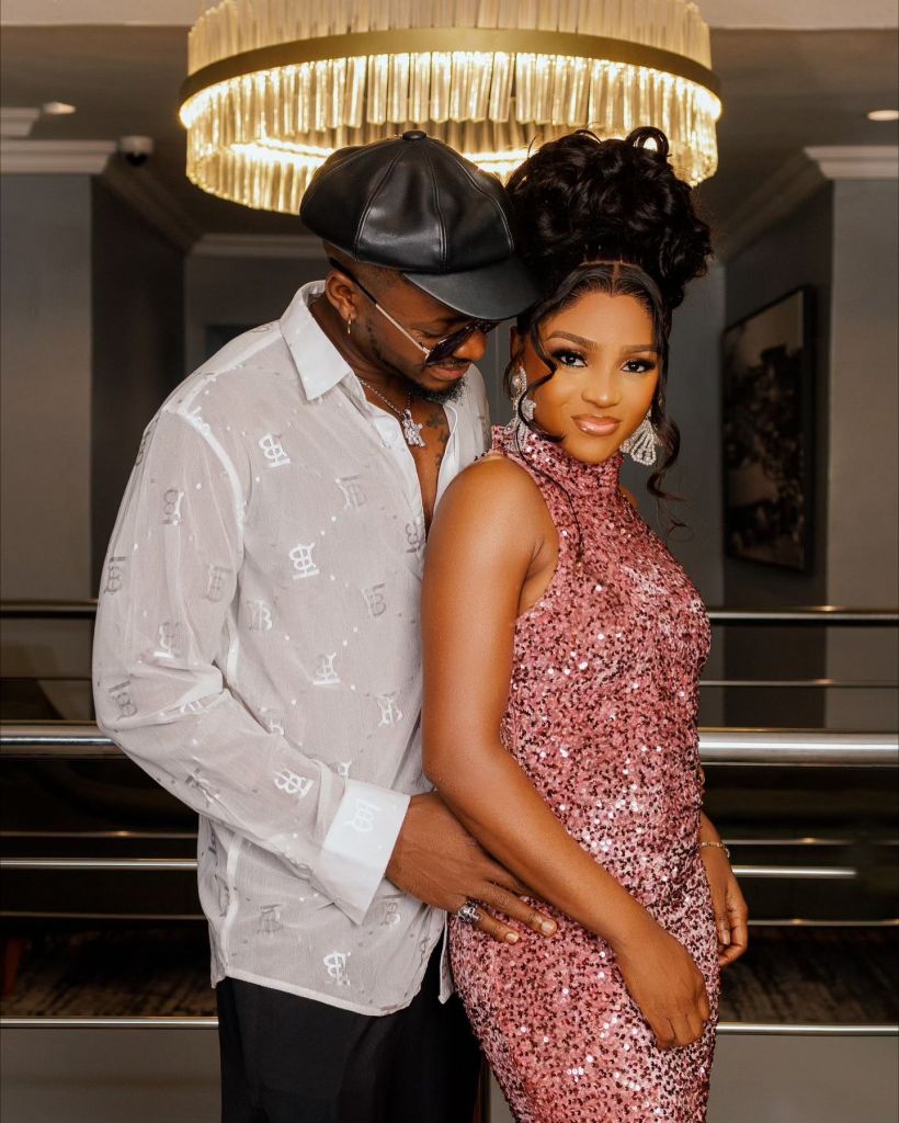 A glamorous BBNaija couple Kellyrae and Kassia stand close together, with Kassia in a sparkling pink dress and Kellyrae in a stylish white shirt and black hat. Their affectionate poses and elegant attire capture a moment of romance and celebration under a chic chandelier.