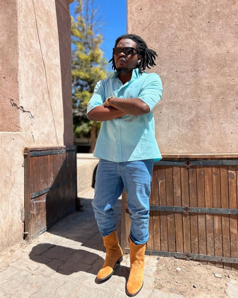 Olamide, on the right, has dreadlocks and wears a light blue shirt, also sporting sunglasses.