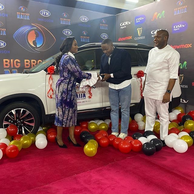 BBNaija season 9 winner Kellyrae celebrates as he receives the keys to his new car from event organizers. Surrounding him are key representatives, with the car adorned in festive ribbons and balloons, highlighting the joyous occasion.