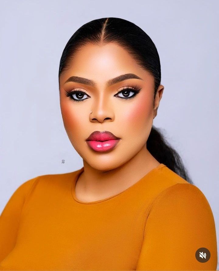 Bobrisky with a smooth, radiant complexion poses confidently, showcasing bold makeup with defined brows, striking eye makeup, and glossy lips. Dressed in a vibrant orange top, the subject exudes elegance and sophistication against a soft, neutral background.