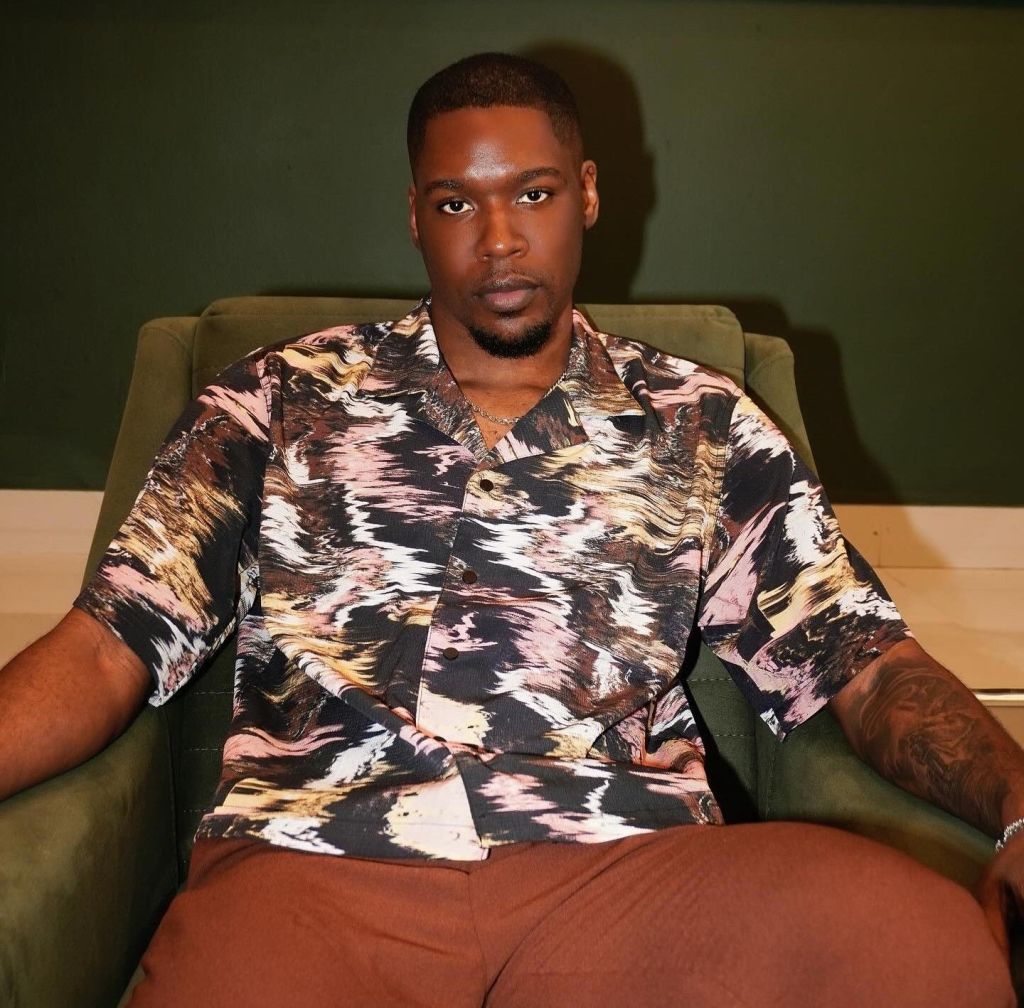 Shaun poses casually in a patterned shirt while sitting on a couch.