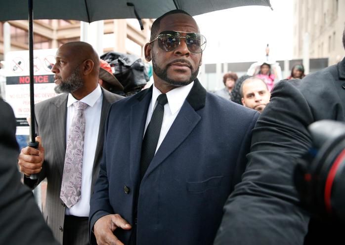 R. Kelly wearing sunglasses and formal attire, standing under an umbrella, amidst a crowd.