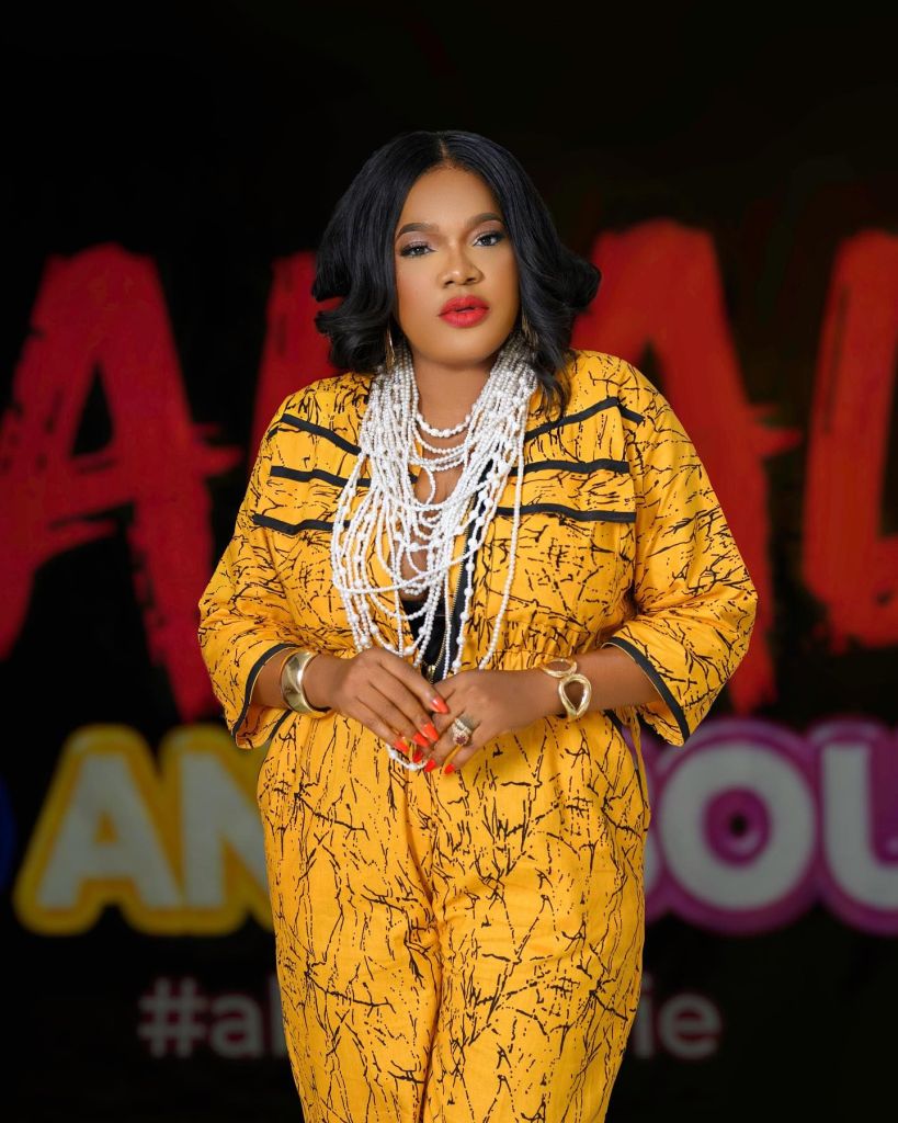 A striking portrait of actress Toyin Abraham wearing a vibrant yellow outfit with a unique black pattern. She accessorizes with multiple strings of pearls and gold jewelry, posing confidently against a dark background with colorful, stylized text that hints at an upcoming project or promotion.
