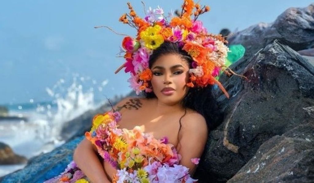 Bobrisky adorned with a vibrant floral crown and a dress made of colorful flowers poses elegantly against a backdrop of rocks and splashing waves in a seaside setting. The scene captures a blend of nature and artistry, emphasizing beauty and creativity.