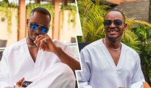 BBNaija star Kellyrae poses confidently in a white outfit, accessorized with sunglasses and jewelry. The backdrop features lush greenery, adding a vibrant touch to the scene. One side captures a playful smirk, while the other displays a warm smile, showcasing a charismatic personality.