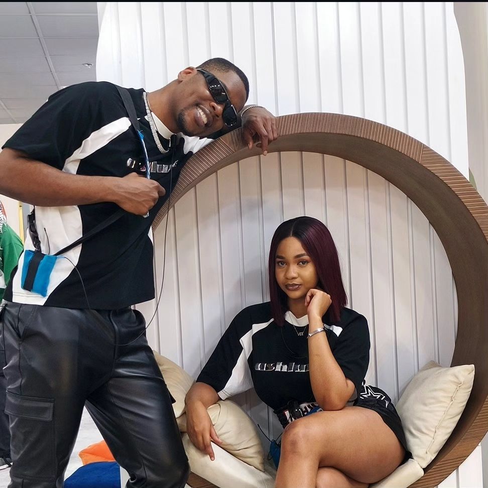 A stylish duo Shaun and Victoria poses together in a cozy setting. Shaun leans playfully against a circular frame, wearing sunglasses and a casual black and white outfit, while the Victoria sits comfortably, showcasing a chic look with long hair and a confident expression. Their relaxed demeanor captures a moment of friendship and fun.