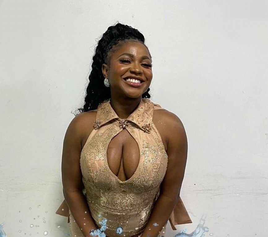 BBNaija star Wanni smiles warmly in a stunning beige dress adorned with intricate embellishments, standing against a plain background. Her hairstyle is curly, and she accessorizes with elegant earrings.