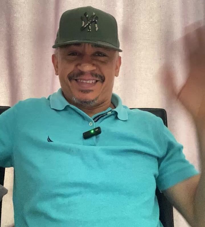Daddy Freeze with a light blue shirt and a cap, smiling at the camera. The background includes colorful text associated with a media or event.