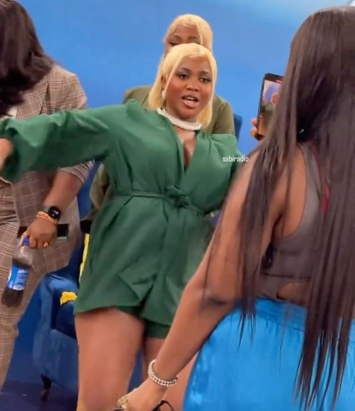 BBNaija Wanni dressed in stylish green outfits, share a joyful moment as they engage with each other, showcasing vibrant expressions. The background features colorful decorations and a vehicle, enhancing the lively atmosphere.