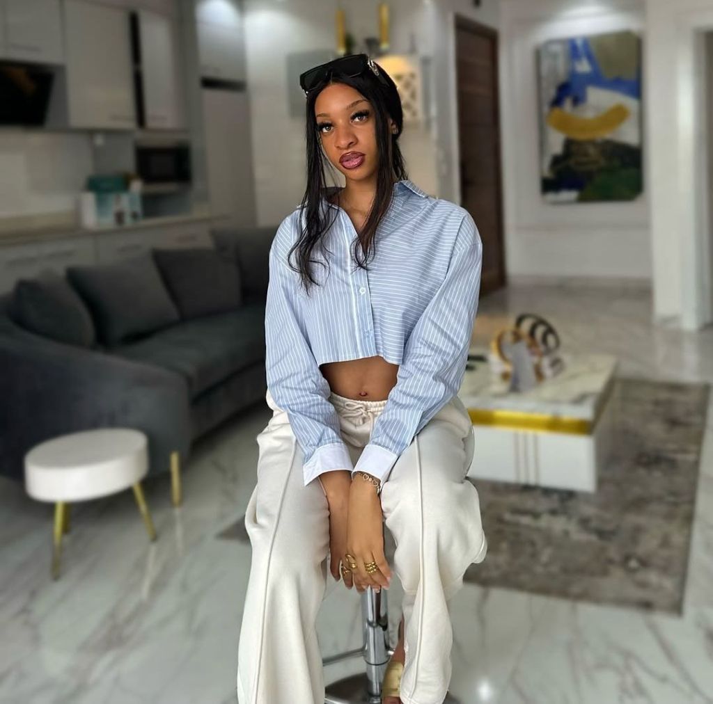 Danielle Edochie in a cropped blue striped shirt and white pants, accessorized with gold rings and sunglasses on her head, sitting on a stool in a modern living room with a grey sofa, white ottoman, and abstract artwork.