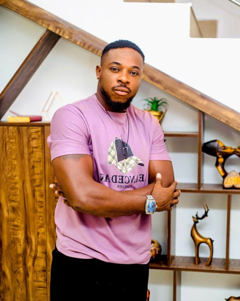 Godwin Nnadiekwe stands confidently in a stylish pink shirt with "LEANCED FOREVER" printed on it, accessorized with a necklace and a watch. He poses with arms crossed against a backdrop of wooden shelves adorned with decorative items, including a stag statue and a potted plant.