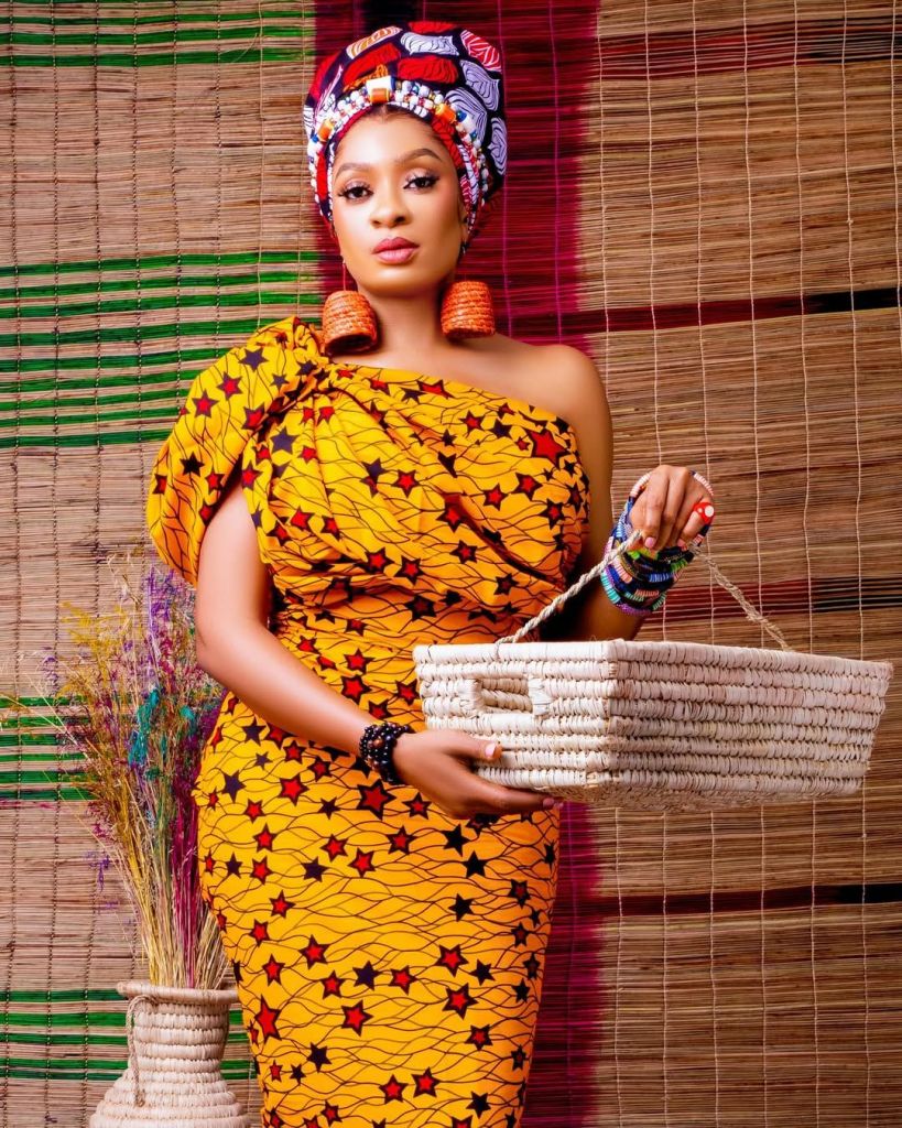 May Edochie in a vibrant yellow dress with black and red star patterns, accessorized with a multicolored headwrap, orange earrings, and a woven clutch, standing against a woven backdrop with green and red accents, holding a white woven basket.