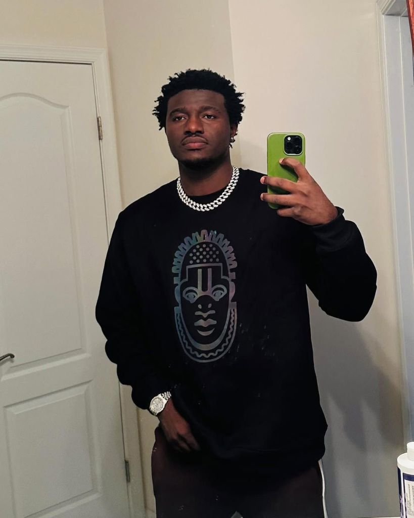 Nasty Blaq is taking a selfie in front of a mirror, wearing a black sweatshirt with a graphic design on it. He has a silver chain around his neck and a watch on his left wrist. The background shows a white door to his left and a white wall behind him.