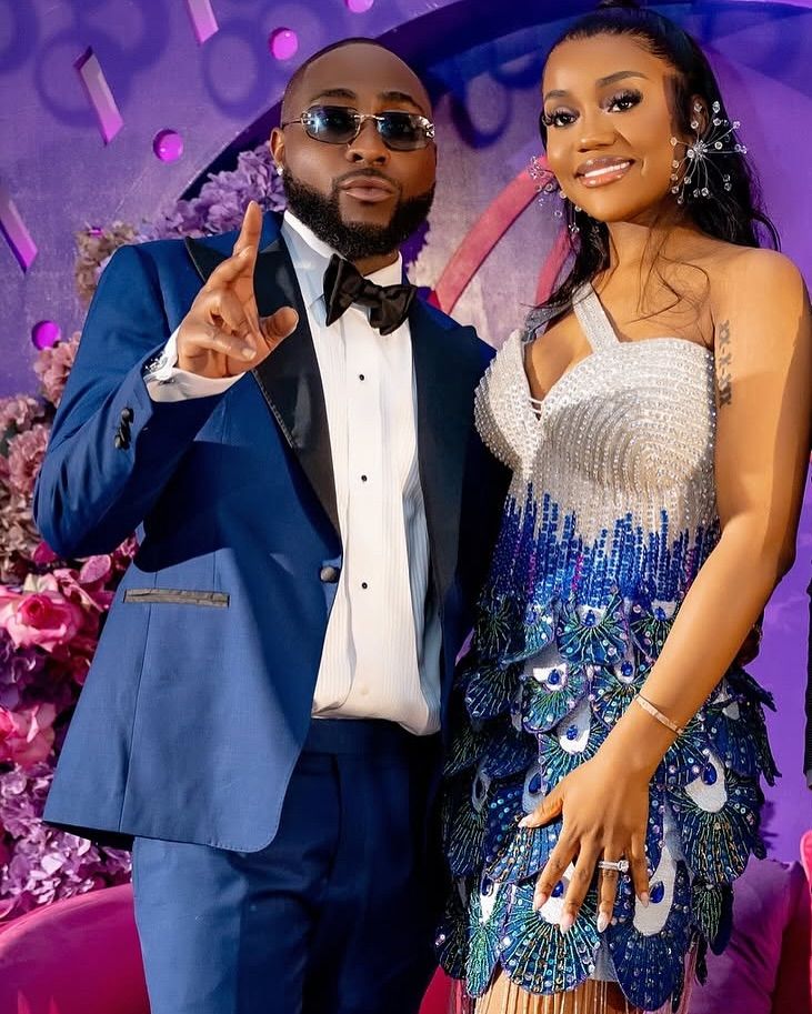 Davido and Chioma pose together at a vibrant event, Davido in a sharp blue suit with a black bow tie, pointing confidently, and Chioma in a stunning silver and blue peacock-feather dress with sparkling earrings and a ring.