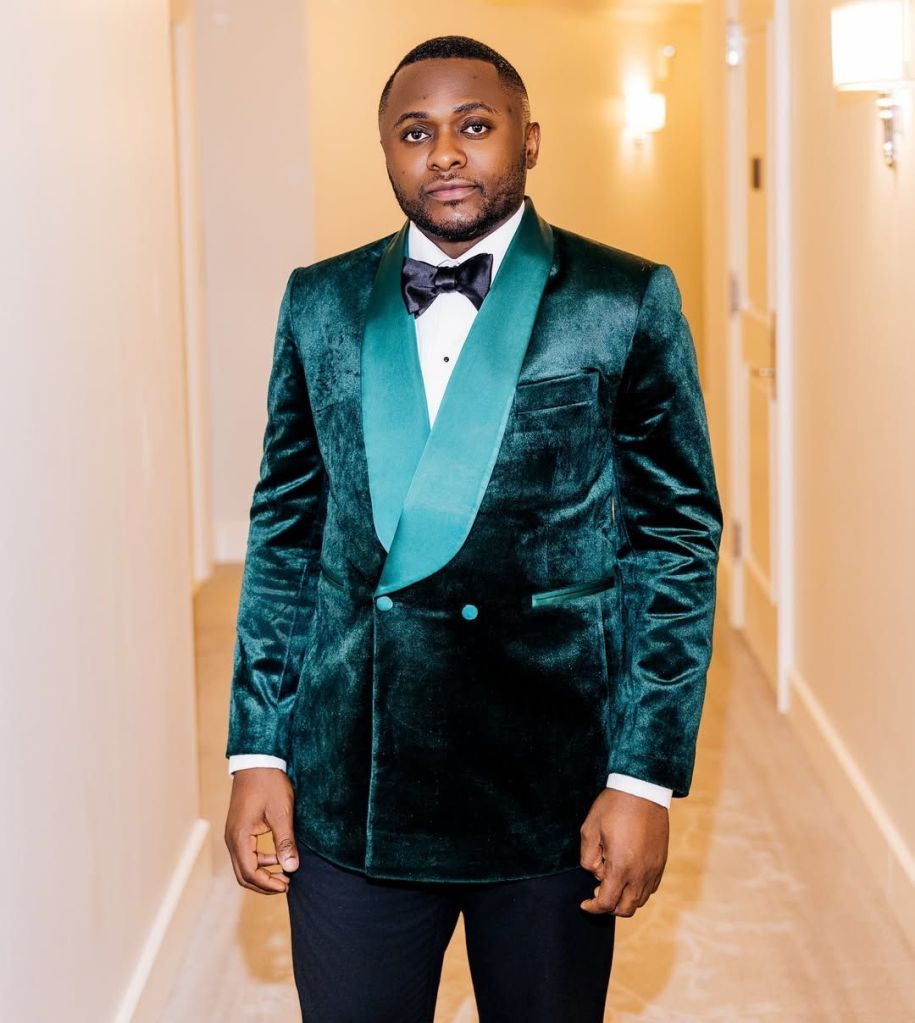 Ubi Franklin in a teal velvet suit with a black bow tie, standing in a warmly lit hallway with wall sconces.