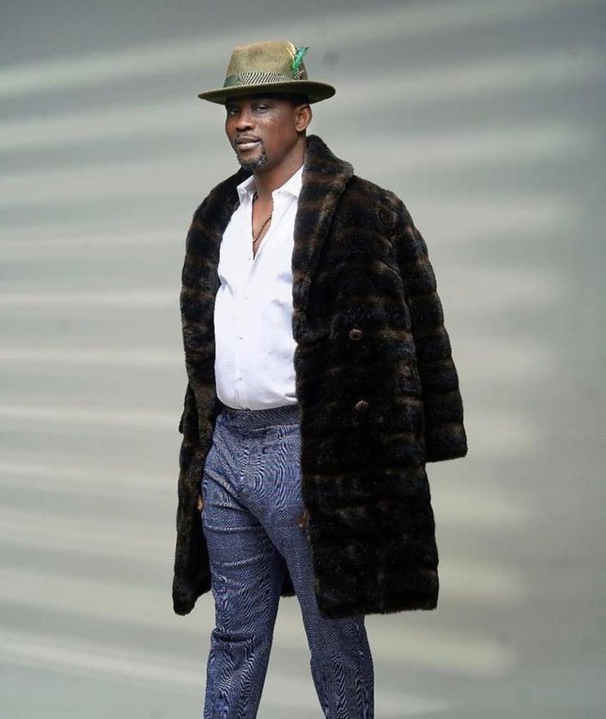 Fuji legend Pasuma stands in a fur coat, white shirt, patterned blue pants, and a green hat with a feather, against a gradient background.
