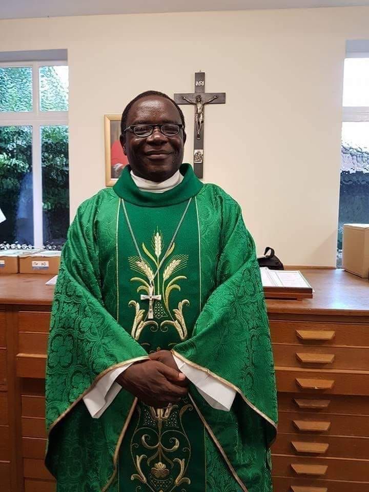Bishop Kukah, wearing a green liturgical vestment with gold embroidery and a pectoral cross, stands with hands clasped in a room with a wooden desk, a crucifix on the wall, and windows in the background.